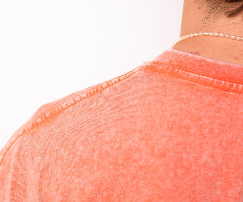 Gros plan sur l’encolure et la couture d’épaule d’un t-shirt corail lavé aux enzymes.